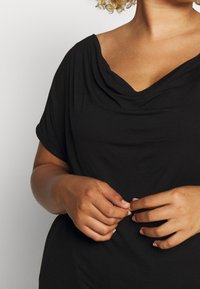 Svart klädd topp med V-ringning, mjukt tyg och korta ärmar; åtsittande runt torso och löst vid fållen för komfort.