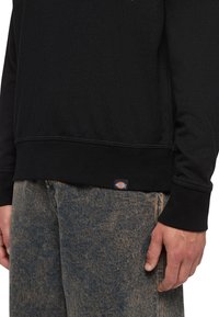 Personne portant un sweatshirt noir avec une petite étiquette logo au-dessus d'un jean délavé à l'acide bleu, la main détendue le long du corps.