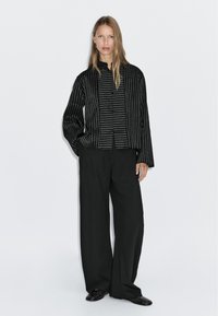 Camicia nera con bottoni, a righe bianche verticali, caratterizzata da un colletto alla cinese e maniche lunghe. Abbinata a pantaloni neri a gamba larga.