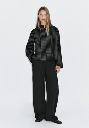 Camicia nera con bottoni, a righe bianche verticali, caratterizzata da un colletto alla cinese e maniche lunghe. Abbinata a pantaloni neri a gamba larga.