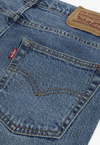 Vaqueros de mezclilla azul con un bolsillo trasero con costuras amarillas, etiqueta roja de Levi's, y un parche de cuero marrón de la marca. Diseño clásico de cinco bolsillos.