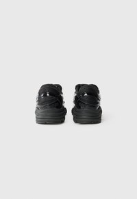 Black athletic shoes with a glossy finish, featuring a padded collar, thick rubber sole, and minimalistic design. Detailed branding on the back.