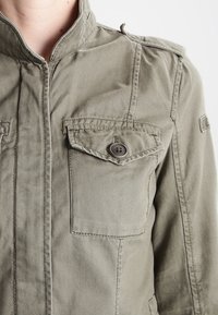 Olivengrøn jakke lavet af bomuld med stående krave, to brystlommer, knaplukning og synlige syningsdetaljer.