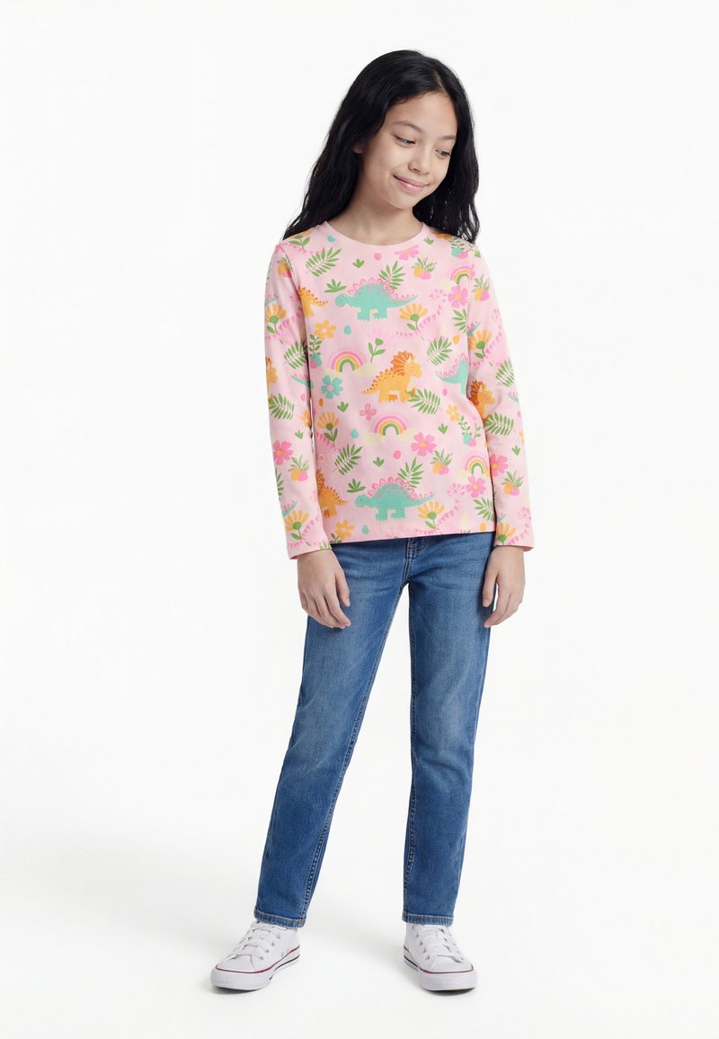 Mergaitė stovi, rankos šalia kūno, vilki rožinį ilgomis rankovėmis marškinėlį su dinozaurais, vaivorykštėmis ir gėlėmis, mėlynas džinsus ir baltus sportinius batelius.