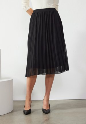 Žena v černé plisované midisukni a černých lodičkách s špičatou špičkou, stojící na betonové podlaze před bílou zdí.