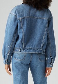 Džínová bunda, světle modrá s bílými skvrnami od barvy, volný střih, knoflíkové manžety a pas. Obsahuje dvě zadní kapsy a akcenty prošití.