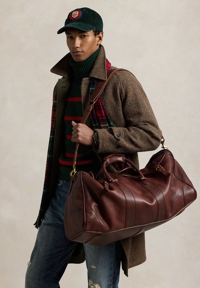 Młody mężczyzna ubrany w brązowy płaszcz, zielony sweter w paski, czapkę i szalik, niosący dużą brązową skórzaną torbę podróżną na ramieniu.