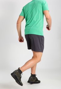 Grön t-shirt med kort ärm, grå shorts och svarta samt olivgrön mönstrade träningsskor med snörning och ankelsupport, sedd från baksidan.