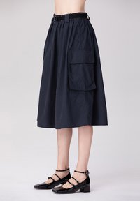 Jupe midi marine avec taille élastique et ceinture, dotée de deux poches latérales. Associée à des chaussures Mary Jane en cuir verni noir avec des brides à la cheville.