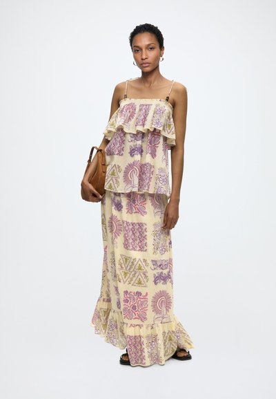 Femme portant une robe longue beige à motifs sans manches avec des volants, tenant un sac à main marron, debout devant un fond blanc uni.