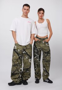 Pantaloni cargo mimetici in verde oliva con motivi a macchie nere e bianche, caratterizzati da una vestibilità rilassata e tasche laterali. Maglietta bianca e canotta.