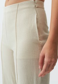 Pantalons beiges taille haute en tissu texturé. Dotés de coutures verticales, d'une fermeture à glissière cachée et d'une coupe droite.