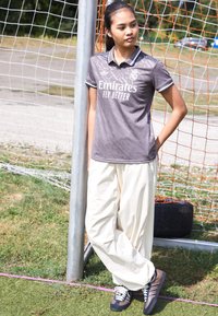 Sivi atletski polo s logom Emirates, uzorak teksture, crna boja ovratnika. Upareno s labavim krem hlačama i crnim tenisicama. Nogometni gol u pozadini.
