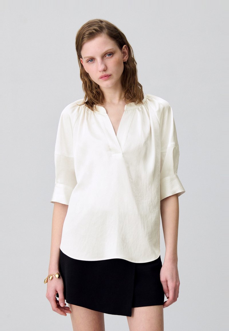 Blusa blanca de manga corta con escote en V y corte relajado, hecha de una tela ligera. Presenta una textura suave y un ligero fruncido en los hombros.