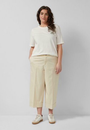 Kobieta o długich, kręconych włosach, ubrana w luźny biały t-shirt, beżowe szerokie spodnie 7/8 oraz białe trampki, stojąca z rękami w kieszeniach.