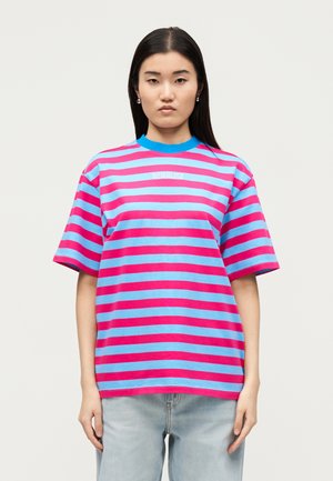 Νεαρή γυναίκα με μακριά μαύρα μαλλιά φοράει φαρδιά ροζ και μπλε ριγέ μπλούζα και ανοιχτόχρωμο μπλε τζιν, στέκεται μπροστά σε ανοιχτό φόντο.