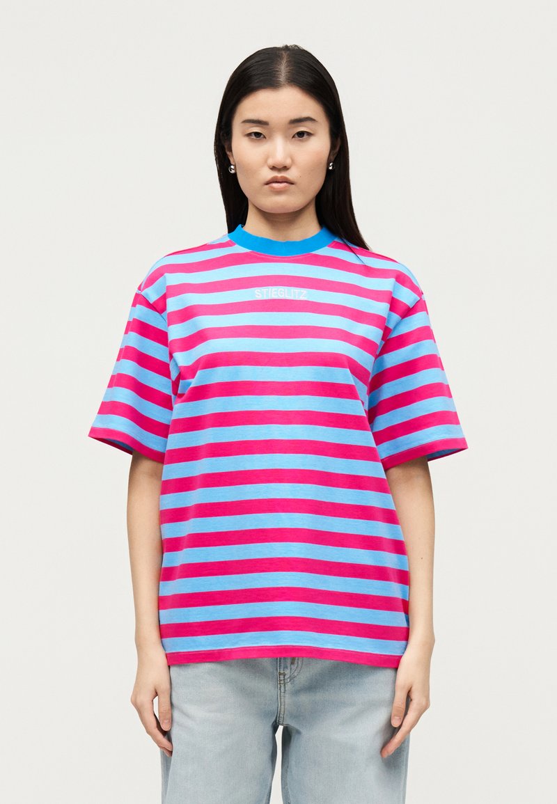 Νεαρή γυναίκα με μακριά μαύρα μαλλιά φοράει φαρδιά ροζ και μπλε ριγέ μπλούζα και ανοιχτόχρωμο μπλε τζιν, στέκεται μπροστά σε ανοιχτό φόντο.