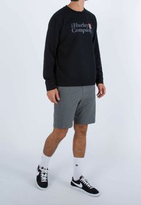 Muž v černé dlouhé mikině Hurley, šedých šortkách, bílých ponožkách do poloviny lýtek a černých teniskách Nike stojí na jednobarevném pozadí.