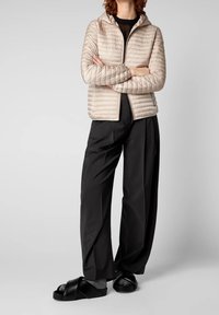 Veste matelassée beige clair avec capuche, fermeture éclair et détails côtelés, associée à un pantalon large noir et des sandales noires à brides croisées.