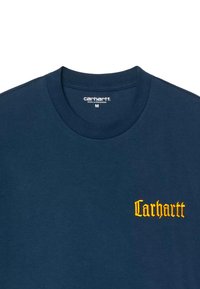 Camiseta de algodón azul marino con cuello redondo; presenta el logo bordado en amarillo de "Carhartt" en el lado izquierdo del pecho; textura suave.