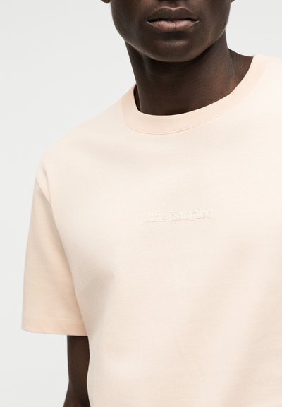 Άνδρας φοράει ανοιχτό μπεζ μπλουζάκι με στρογγυλή λαιμόκοψη και κοντά μανίκια, με λευκό κεντημένο λογότυπο "The Kooples" στο στήθος.