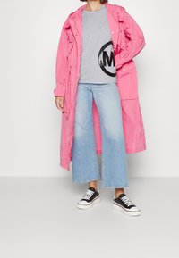 Lange roze regenjas, grijs T-shirt met zwart logo, lichtblauwe wijde jeans en zwarte sneakers met witte zolen.