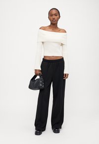 Rippoptik, schulterfreies weißes Crop-Top kombiniert mit schwarzen weit geschnittenen Hosen, hält eine kleine schwarze Tasche mit Ösen-Details. Glatte Textur.