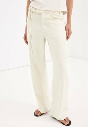 Personne portant un pantalon blanc ample taille haute avec une ceinture à nouer sur le côté et des sandales noires type tongs sur un sol blanc.