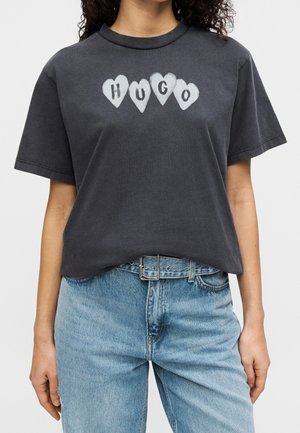 Femme portant un t-shirt gris foncé avec "HUGO" en formes de cœur blanches sur la poitrine et un jean taille haute bleu clair avec une ceinture noire.