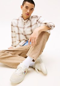 Zapatillas beige con tejido texturizado, cordones blancos y un pequeño detalle de logotipo. El modelo lleva una camisa de cuadros abotonada y pantalones marrón claro.