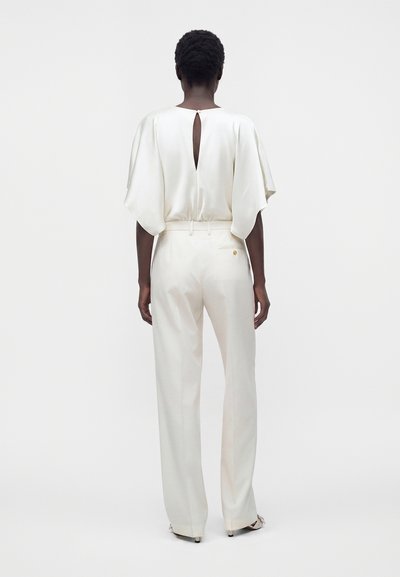 Personne aux cheveux courts et bouclés porte un pantalon blanc à jambes larges et un haut blanc ample avec une ouverture en forme de goutte au dos, debout devant un fond uni.