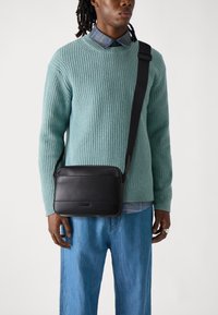 Sort lærremseveske med glatt tekstur, med et flatt design og justerbar stropp, båret over blå denim. Kraftig tegrønn strikkegenser.