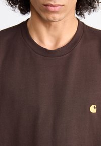 T-shirt in cotone marrone scuro con colletto tondo e maniche corte, caratterizzato da un piccolo logo giallo in basso a destra. Texture liscia.