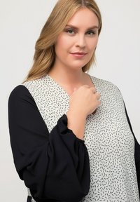 Blusa con mangas negras y cuerpo blanco que presenta un patrón negro disperso. Diseño con escote en V y mangas sueltas y fruncidas.