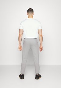 Man die van achteren gezien wordt, gekleed in een wit shirt met korte mouwen, lichtgrijze joggerbroek en zwarte sneakers, tegen een effen achtergrond.