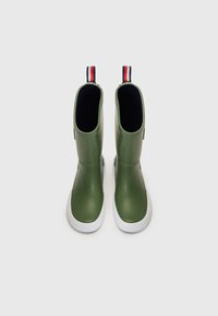Bottes de pluie en caoutchouc vert avec une texture lisse, semelles blanches et une languette rayée rouge, blanche et bleue en haut.