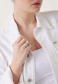 Anello in argento con finitura testurizzata e piccolo accento a forma di cuore, indossato su una mano che tiene il colletto di una giacca in denim bianca. Top bianco visibile sotto.
