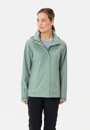 Veste imperméable vert clair avec capuche, dotée de fermetures éclair et de Velcro, portée sur un haut bleu, associée à un pantalon sombre.