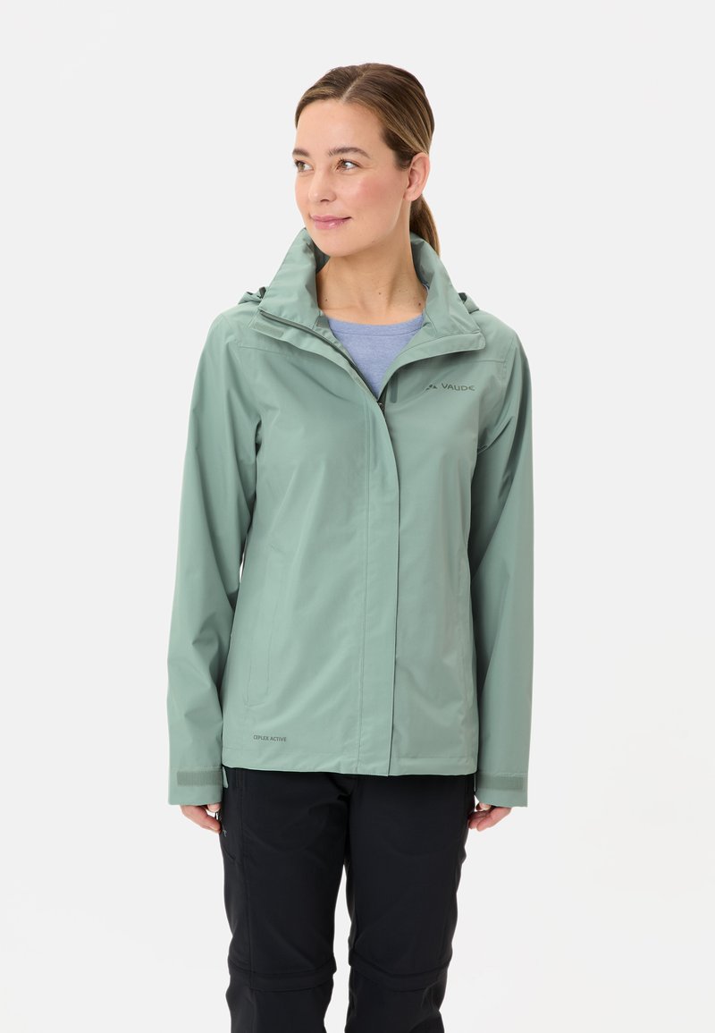 Veste imperméable vert clair avec capuche, dotée de fermetures éclair et de Velcro, portée sur un haut bleu, associée à un pantalon sombre.