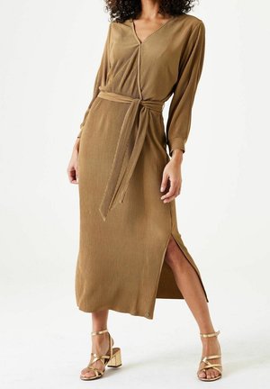 Vrouw draagt een langarmige, geribbelde midi-jurk in camelkleur met een zijsplit en bijpassende riem, gecombineerd met gouden sandalen met blokhak en bandjes.