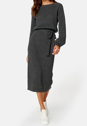 Kobieta w ciemnoszarym prążkowanym swetrze i dopasowanej midi spódnicy z wiązaniem w talii, w białych trampkach, idąca do przodu.