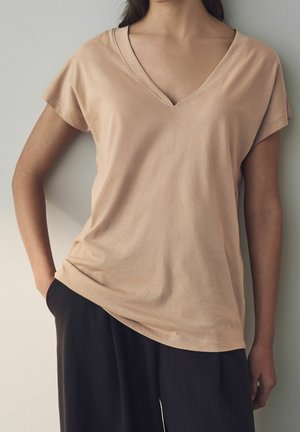 Nainen, jolla on beige V-kaula-aukollinen lyhythihainen paita ja jolla on toinen käsi taskussa, seisoo vaaleaa seinää vasten, näkyvissä ylävartalo ja rintakehä.