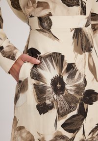 Robe en tissu crème avec de grands motifs floraux marron, dotée d'une taille cintrée et d'une poche latérale. Texture lisse et manches longues visibles.