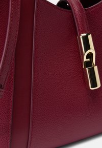 Bolsa de couro burdeos com acabamento texturizado, apresentando um fecho de metal dourado e uma alça de ombro resistente. Linhas limpas e hardware minimalista.