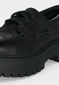 Mocassin en cuir noir avec motif texturé, semelle épaisse à crampons, fermeture à lacets sur l’avant, et logo arbre embossé sur la partie latérale.