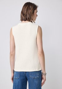 Ärmeløs cremet sweater med ribbet tekstur, der har en rund halsudskæring og lige kant, båret med blå højtaljede jeans.