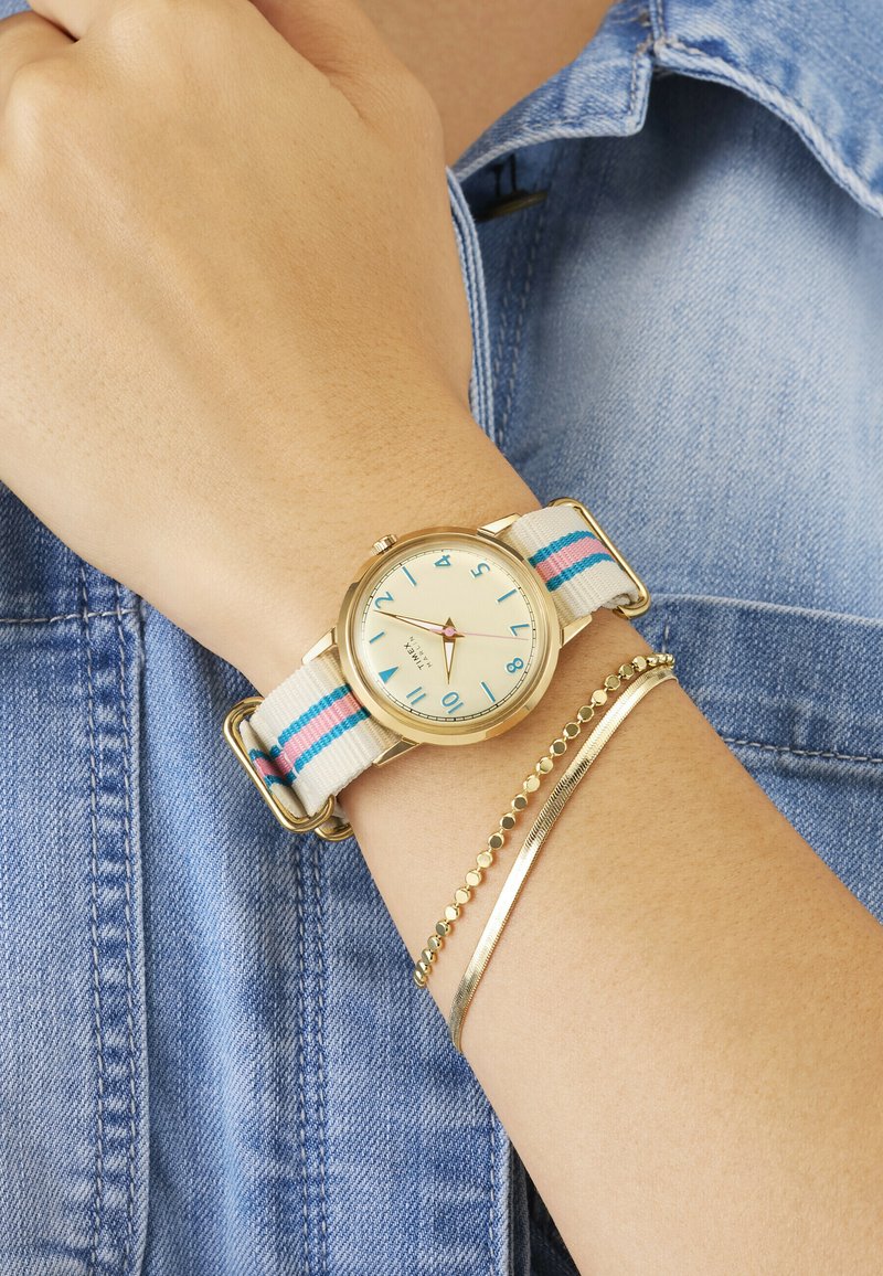 Poignet avec une montre à boîtier en or, cadran crème avec chiffres bleus, bracelet en tissu rayé, et deux bracelets délicats en or, porté sur une veste en jean.