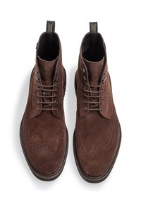 Stivaletti alla caviglia in suede marrone con design brogue, dotati di lacci tondi e di una linguetta sul tallone, su uno sfondo bianco.