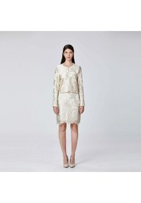 Femme portant un tailleur à jupe en dentelle florale crème avec des manches longues et des talons assortis, se tenant devant un fond clair.