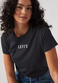 Melna T-kreklu ar īsām piedurknēm un apaļu kakla izgriezumu, ar balto "LEVI'S" logotipu uz krūtīm, apvieno ar ziliem džinsiem.